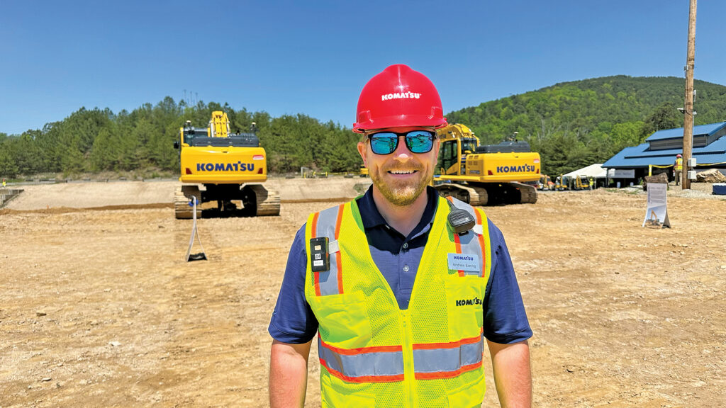 A man wearing a red hard hat and a Komatsu reflective vest, identified as Andrew Earing, Director of Operator and Technical Training at Komatsu, standing in front of two Komatsu excavators.
