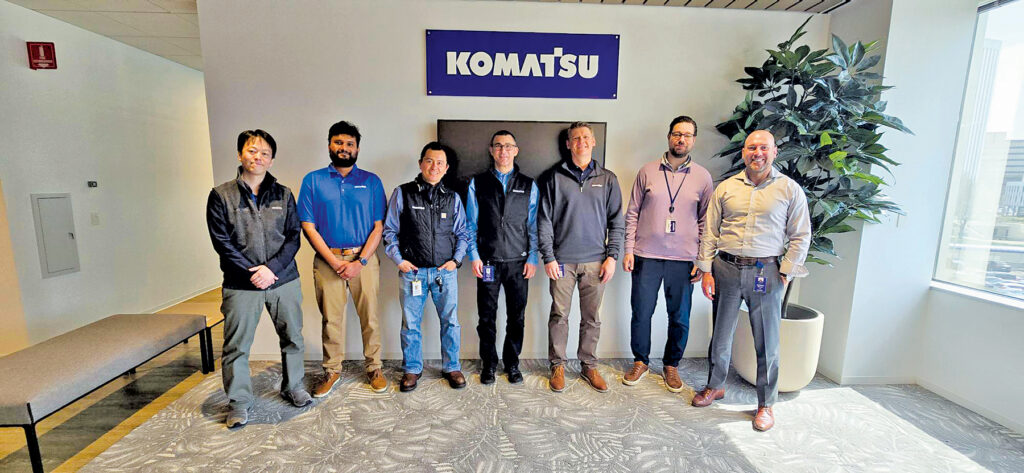 Seven men standing in an office setting in front of a Komatsu sign. From left to right: Eric Uehara, Krutarth Desai, Isamu Hamai, Goran Zeravica, Arek Krynski, Pierre Deering, and Matt Beinlich, members of Komatsu's remanufacturing team.
