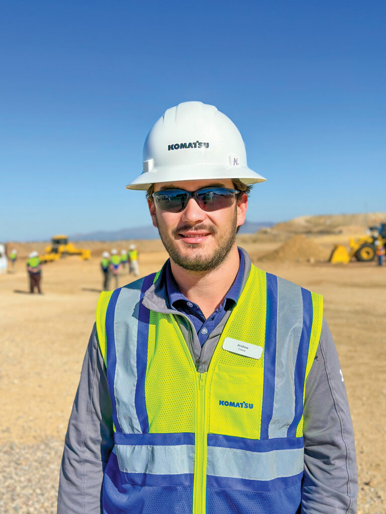 An image of Andrew Casey, Digital Solutions Analyst at Komatsu, wearing a white Komatsu hard hat and reflective vest, standing a distance away from Komatsu machines and other people behind him.