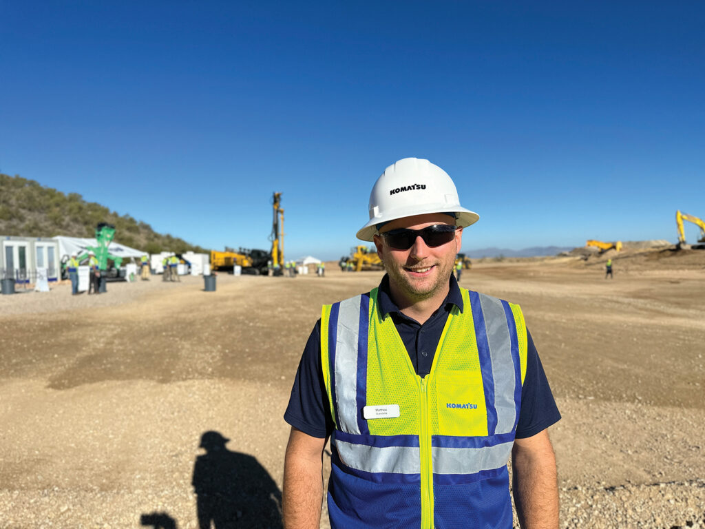 An image of Matt Buerstetta, Product Manager at Komatsu, wearing a white Komatsu hard hat and reflective vest, and sunglasses, standing a distance away from Komatsu machines and other people behind him.