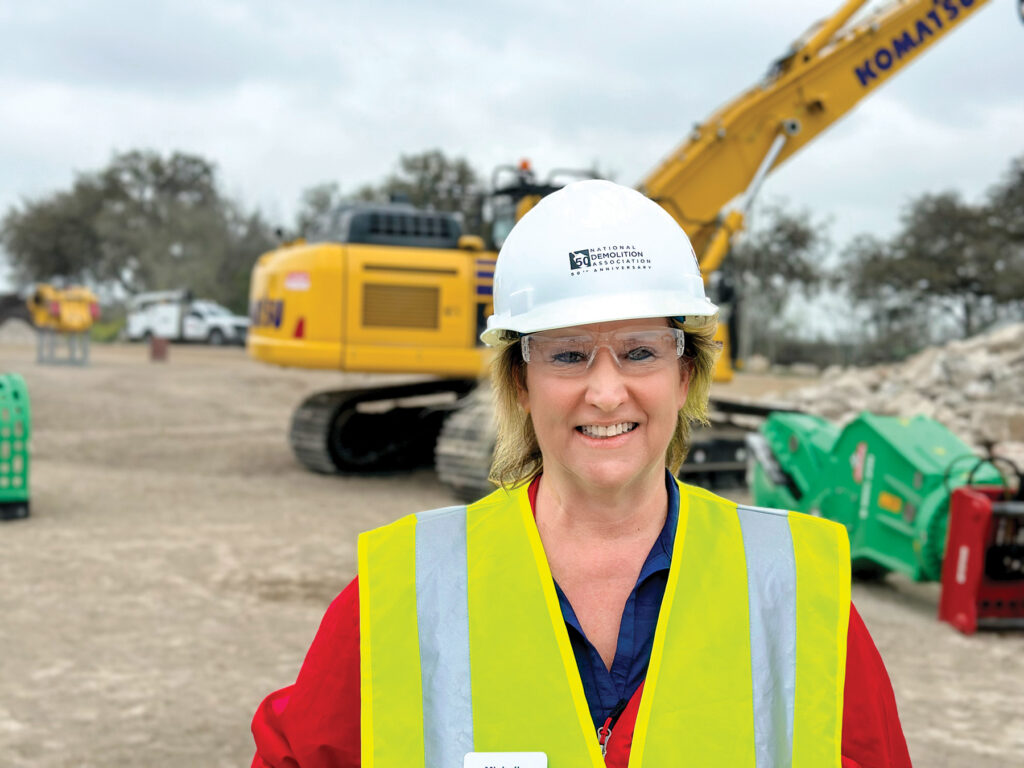 Michelle Wilson, Sales Engineer at Montabert, standing in front of a Komatsu excavator wearing a National Demolition Association 50th anniversary hard hat and a reflective vest.