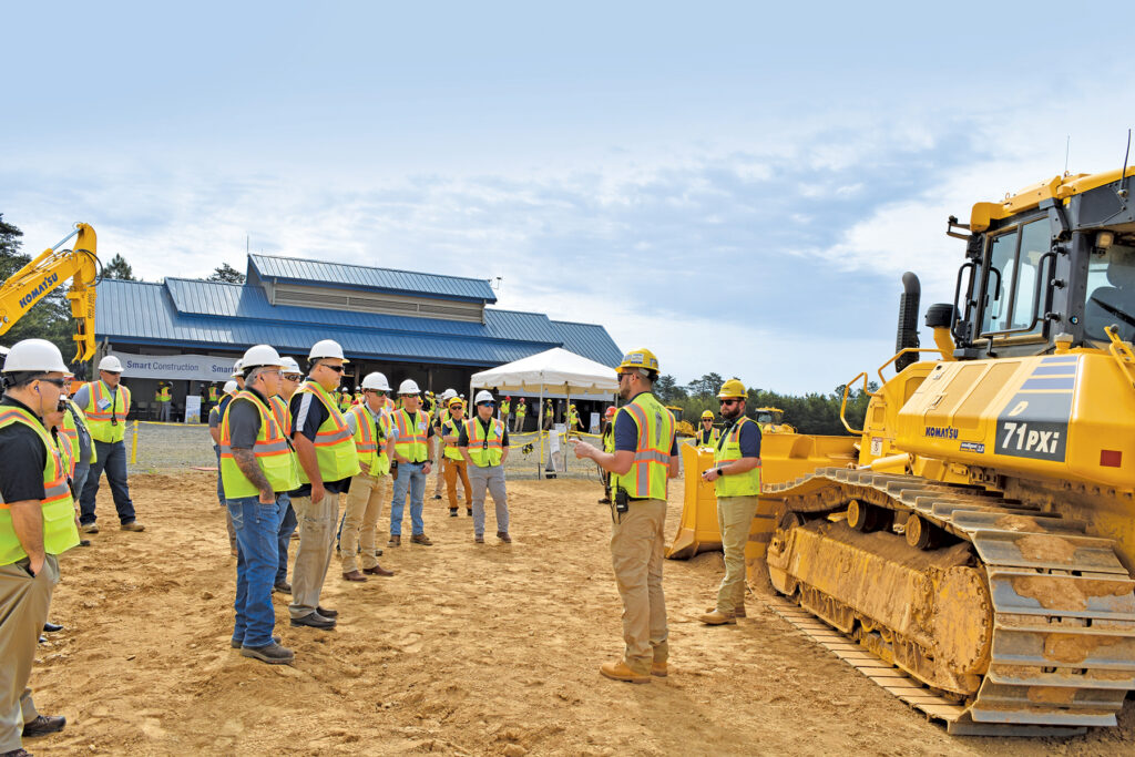 Two men wearing yellow hard hats and reflective vests engage with a crowd of individuals wearing white hard hats and reflective vests. They stand in front of a Komatsu D71PXi bulldozer.