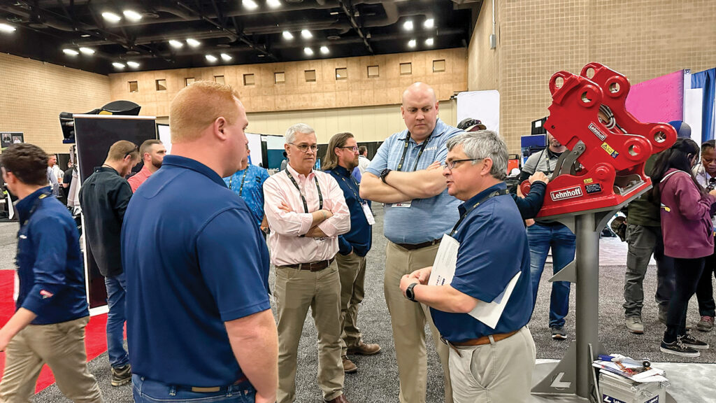 A group of men in work clothes engaged in conversation with each other in an indoor convention setting.