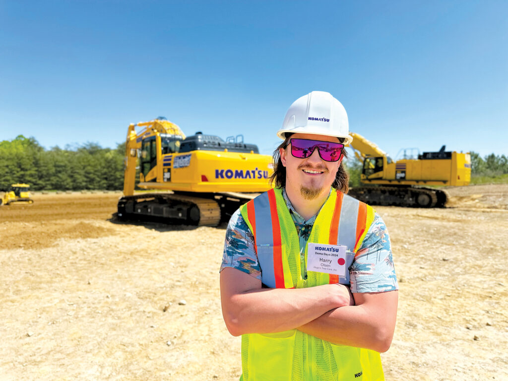 Harry Olsen, COO/Co-owner of Hugo Tree, wearing a Komatsu hard hat and reflective vest while standing in front of Komatsu excavators.
